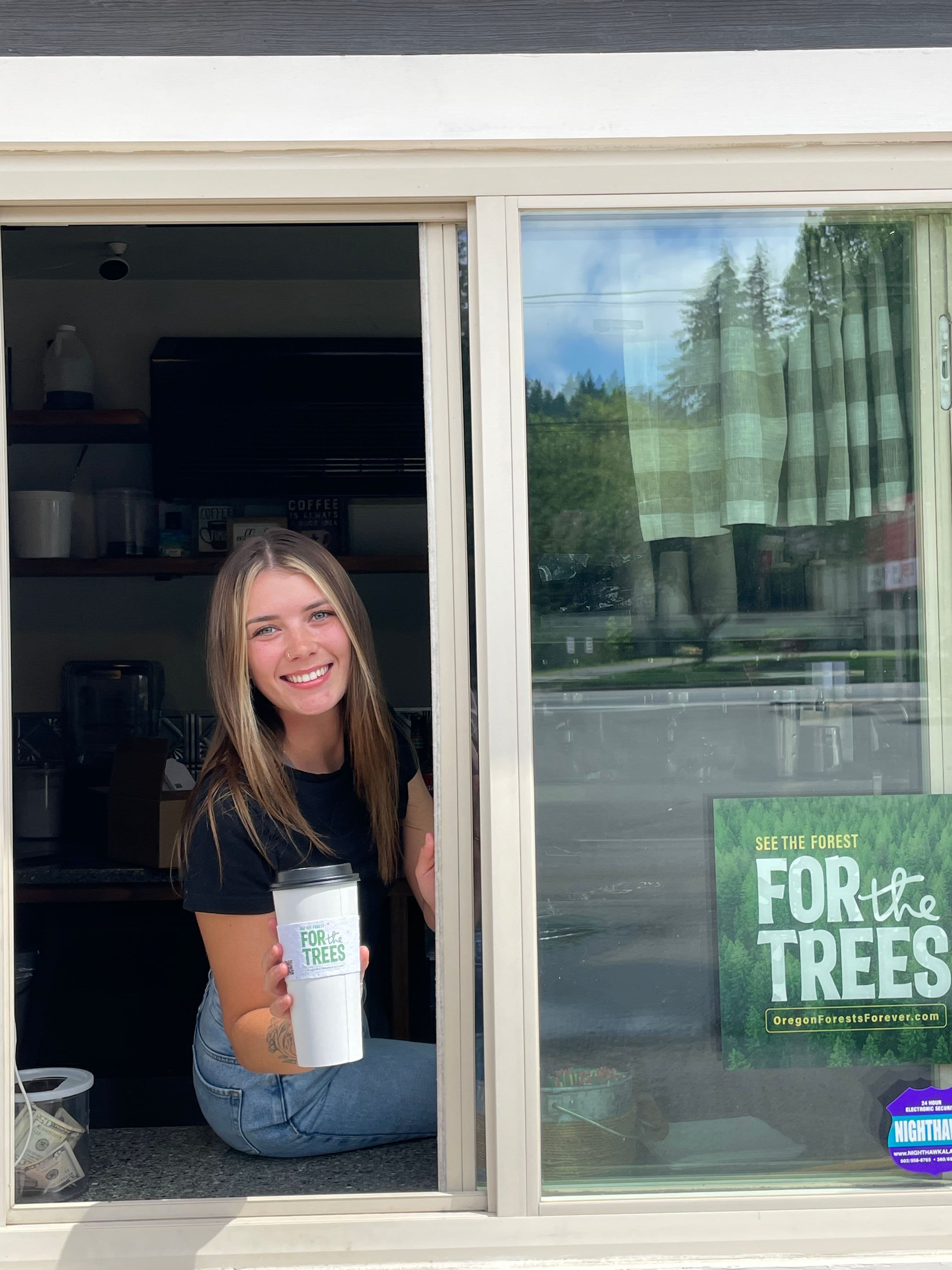 A barista holding a branded coffee cup at a coffee shop window displaying a campaign window graphic
