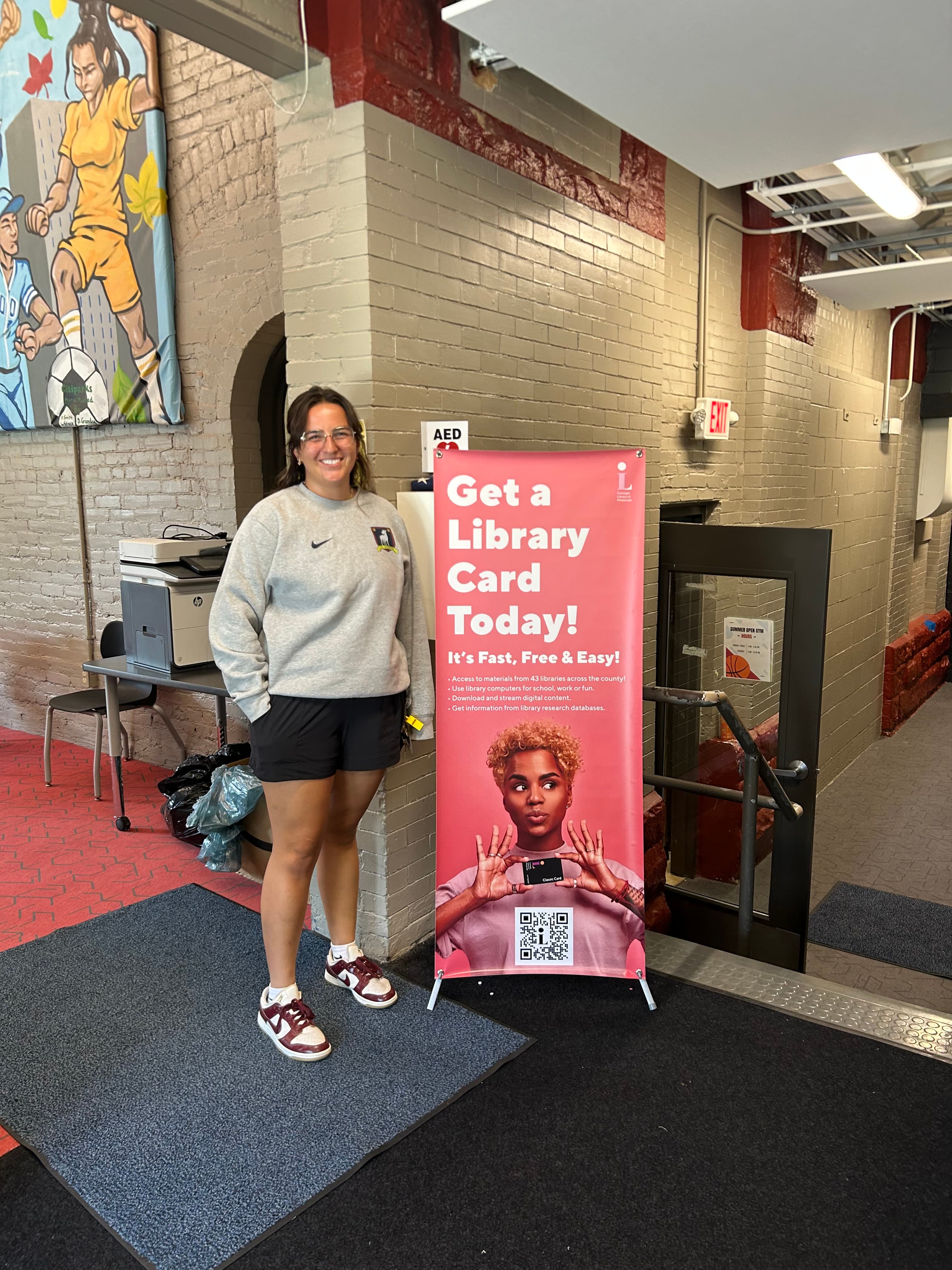 A staff member standing beside a library-card campaign banner inside a recreation center entryway